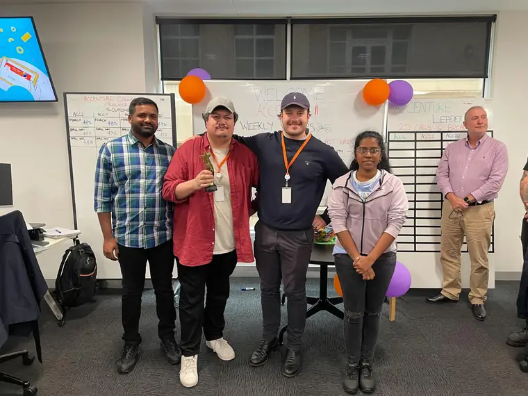 Four colleagues standing together in a contact centre, one holding a small star trophy, with whiteboards and balloons behind them